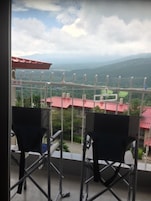 A balcony with two chairs and a metal railing, overlooking a landscape with red-roofed buildings and green hills.