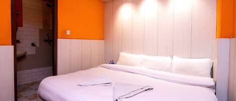Una habitación de hotel con cama, ropa de cama blanca, zona de ducha y paredes anaranjadas.