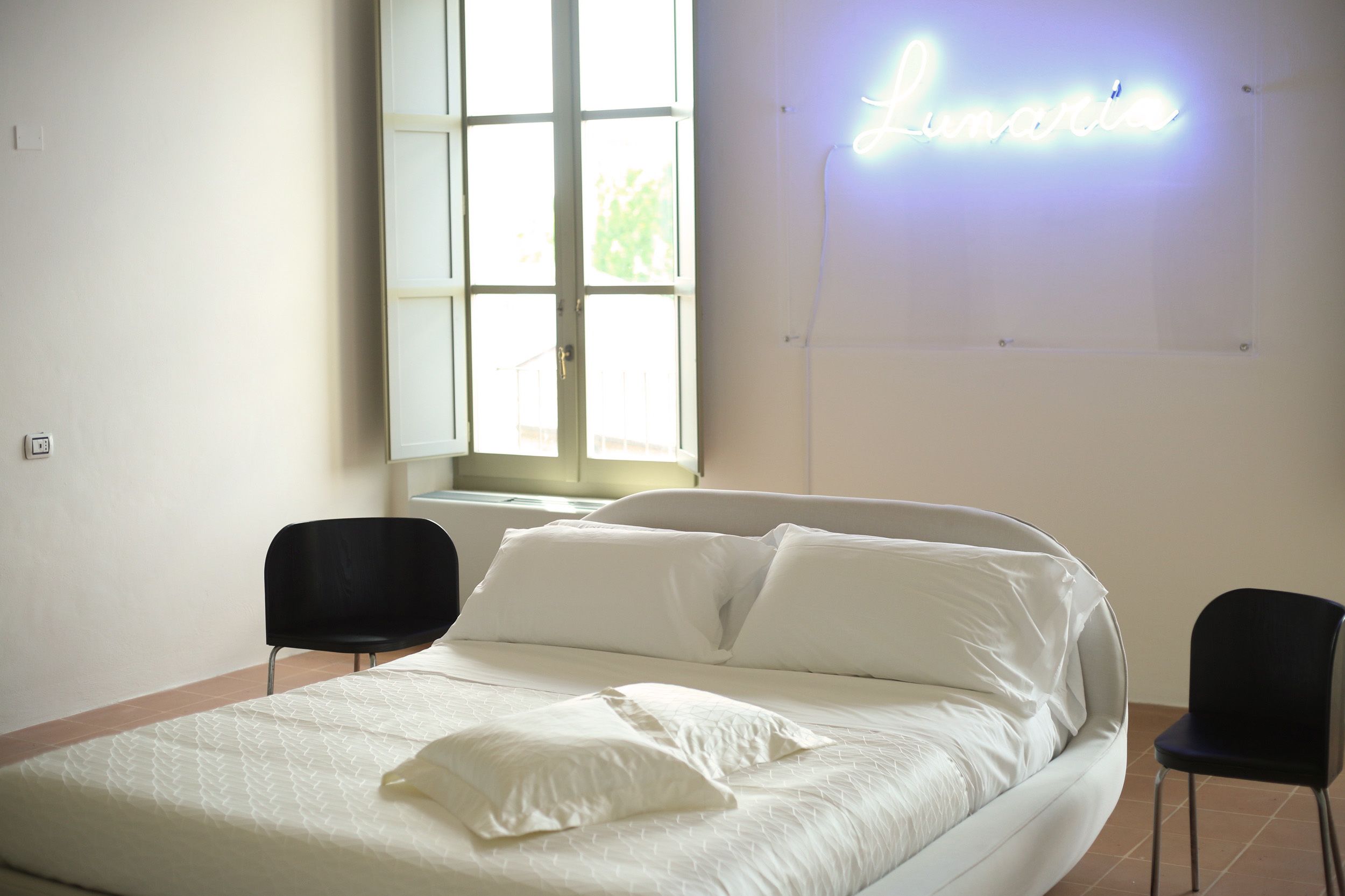 Une chambre d’hôtel avec un lit, deux chaises noires, une grande fenêtre et un panneau lumineux au néon sur le mur.