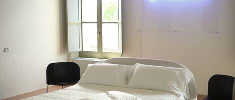 A hotel room with a bed, two black chairs, a large window, and a neon sign on the wall.