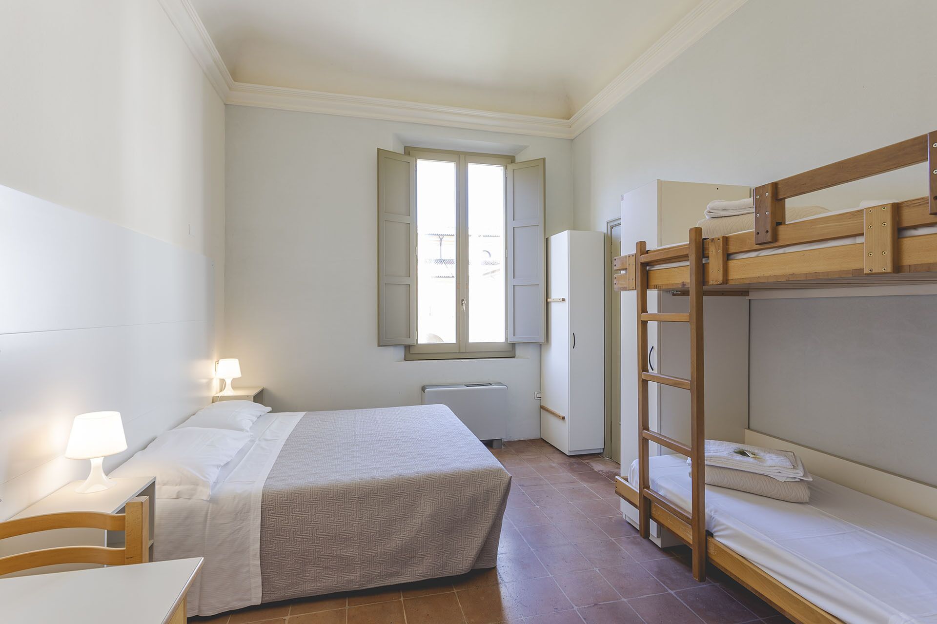Quarto compacto com uma cama beliche, uma cama de solteiro, uma escrivaninha com luminária, um radiador e uma janela com persianas.
