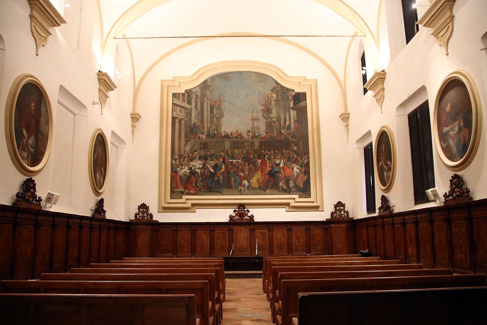 A church interior with wooden pews, a large central painting, and framed artworks on the walls.