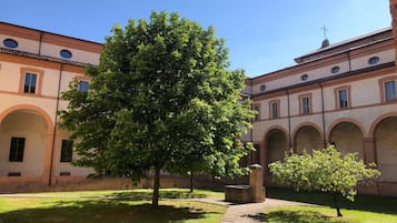 Une cour avec un grand arbre, un chemin en briques et un bâtiment doté d’arcades et d’une croix sur le toit.