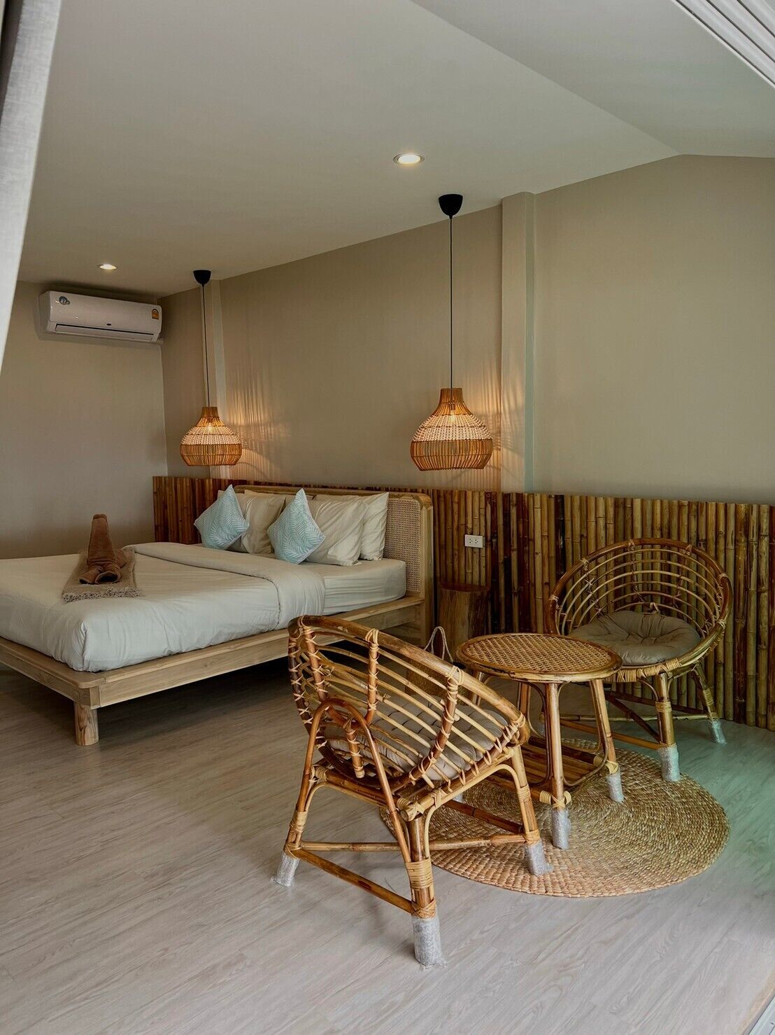 Een kamer met een bed, twee rieten stoelen, een klein rond tafeltje en twee hangende rieten lampen.