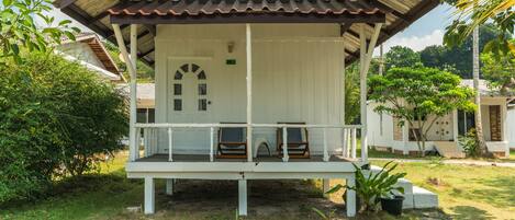 A white building with a brown roof, a porch with two wooden chairs, and a door with a window.