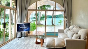 Een ruime woonkamer met een grote televisie, comfortabele zitplaatsen en uitzicht op een tropisch strand en de oceaan.
