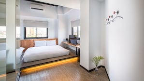 Une chambre d’hôtel moderne, équipée d’un lit, d’un bureau avec un ordinateur, d’une petite plante et d’une fenêtre donnant sur la ville.