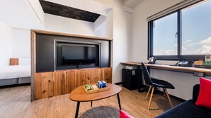Une chambre moderne avec un téléviseur fixé sur un panneau en bois, un bureau avec une chaise, une petite table avec des collations et une grande fenêtre donnant sur le ciel.