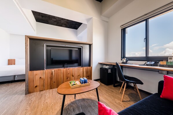 Une chambre moderne avec un téléviseur fixé sur un panneau en bois, un bureau avec une chaise, une petite table avec des collations et une grande fenêtre donnant sur le ciel.