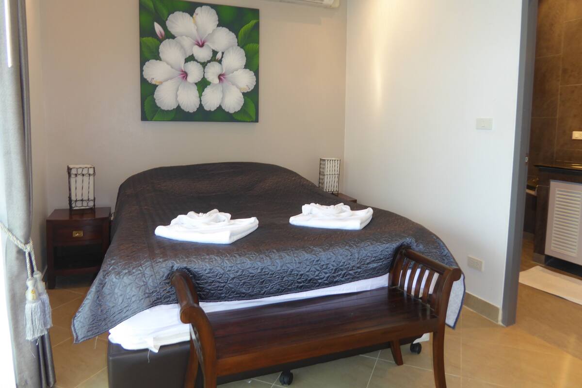A hotel room with a bed, a bench, a painting of flowers, and an air conditioning unit.