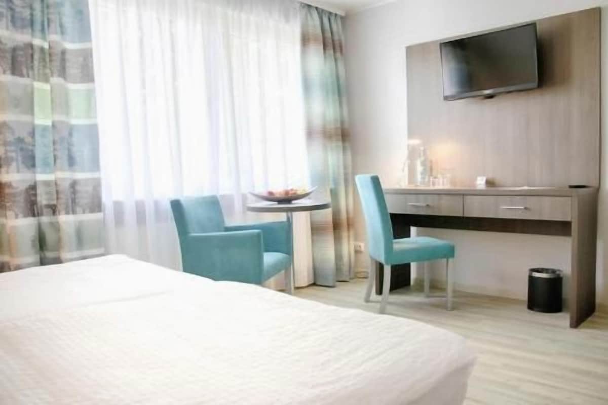 Habitación de hotel con cama, escritorio, silla, televisor y ventana con cortinas.