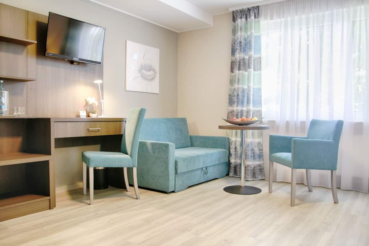 Een moderne woonkamer met een bank, twee fauteuils, een klein tafeltje, een aan de muur bevestigde televisie en een raam met dunne gordijnen.