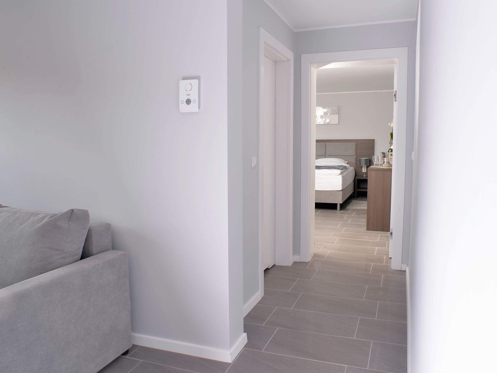 A modern, minimalist living room with a grey sofa and a hallway leading to a bedroom with a bed and bedside table.