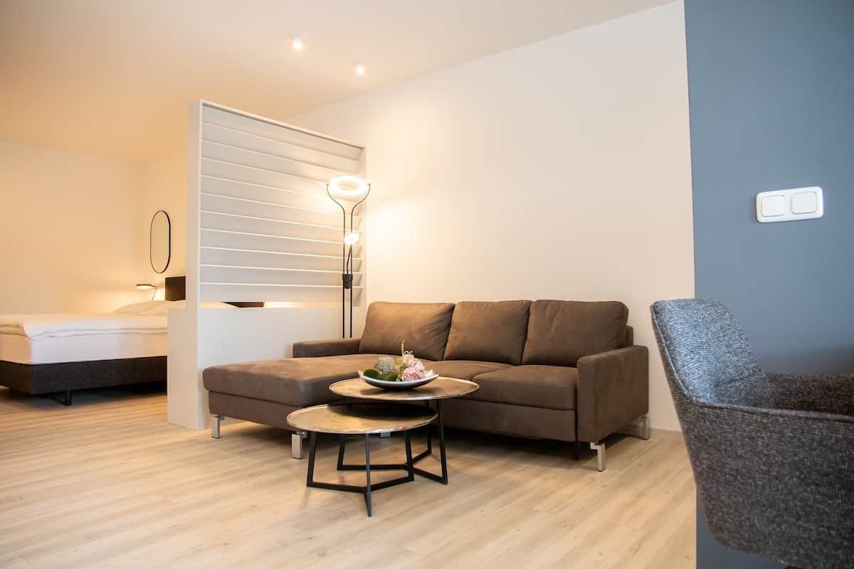 Een moderne woonkamer met een bank, een ronde salontafel en een stoel. Achtergrond toont een bed met een witte hoofdbord.
