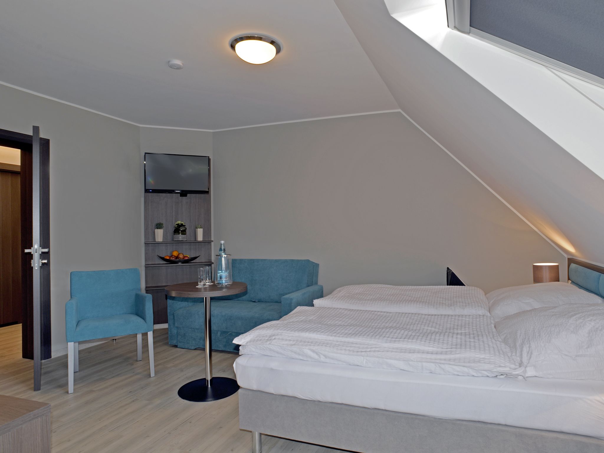 Una habitación moderna y luminosa con una cama, un sillón azul, una mesita con una botella de agua y un televisor montado en la pared.