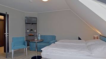 Una habitación moderna y luminosa con una cama, un sillón azul, una mesita con una botella de agua y un televisor montado en la pared.