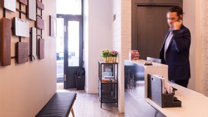 A reception area with a bench, a wall with framed artwork, a desk with a computer and a phone, and a door leading outside. - Hotel Terre Neuve (Paris)