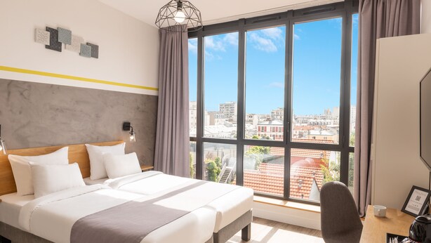 A hotel room with a large bed, a desk, and a view of the city through large windows. - Hotel Terre Neuve (Paris)