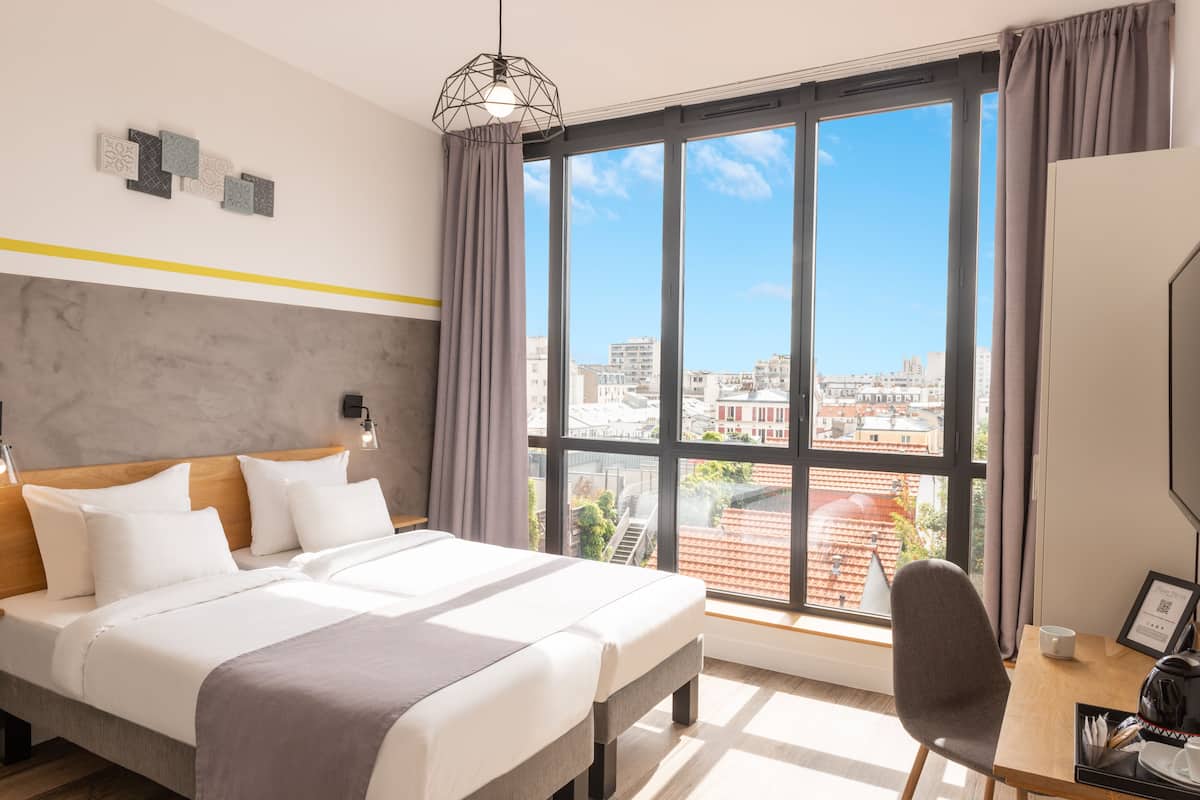 A hotel room with a large bed, a desk, and a view of the city through large windows.