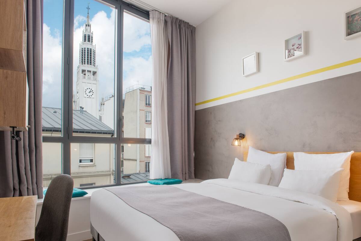 A hotel room with a large window, a bed with white bedding, a desk, and a chair.
