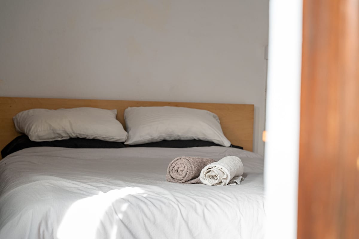 Un lit bien fait, avec du linge de lit blanc et deux serviettes enroulées, une tête de lit en bois et un mur blanc sobre.