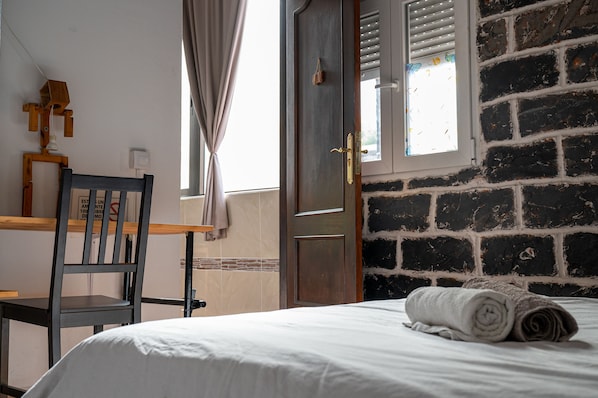 Una habitación de hotel con una cama, una silla de madera, una ventana con persianas y una pared de ladrillo.