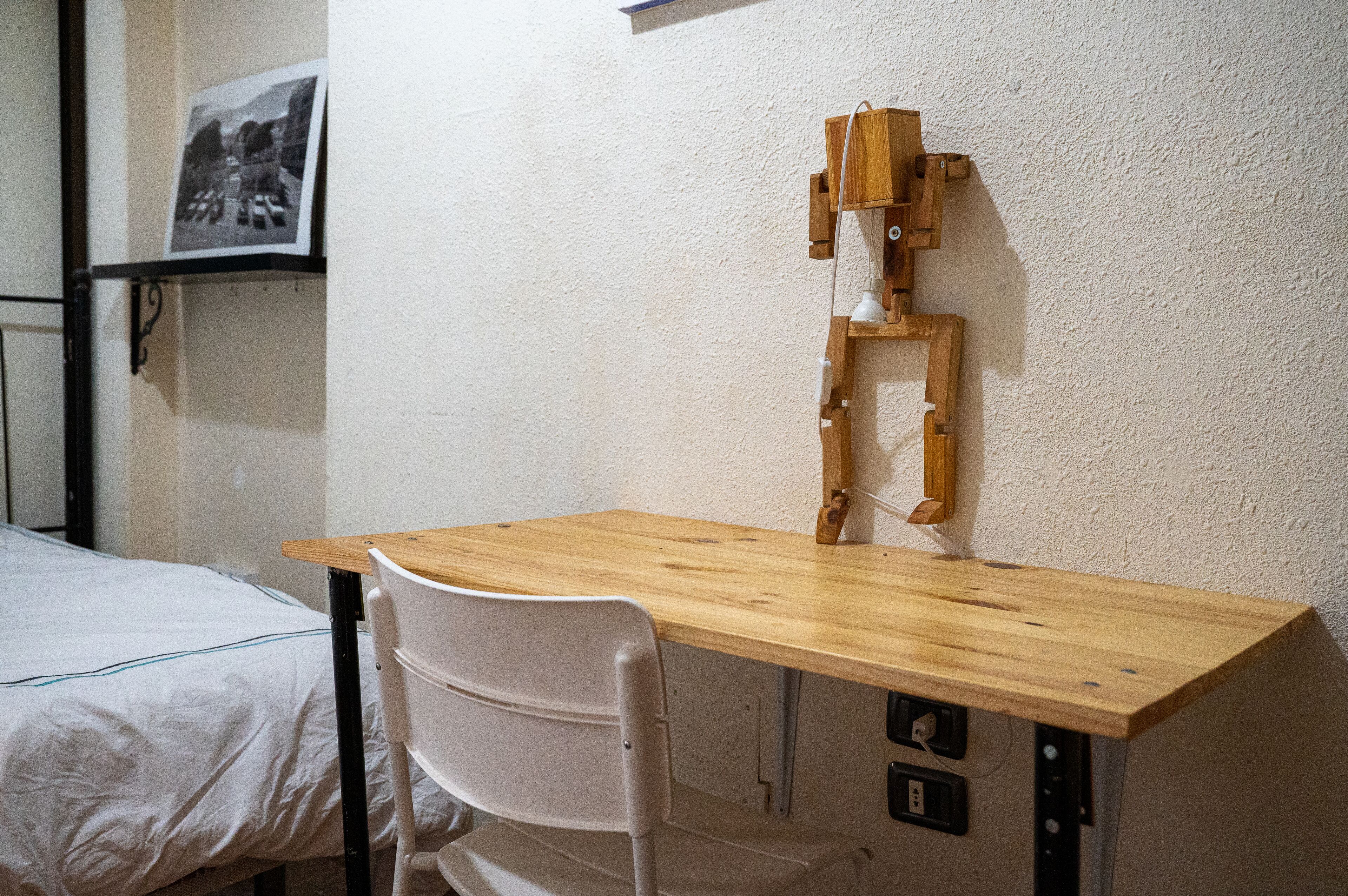 Un escritorio de madera con una silla, una cama y una lámpara fijada a la pared.