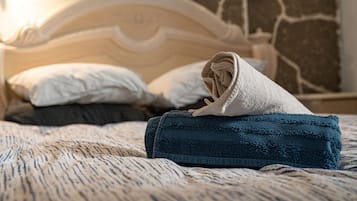 Una cama cuidadosamente tendida con una toalla doblada y un edredón enrollado, un cabecero blanco y una pared de piedra al fondo.