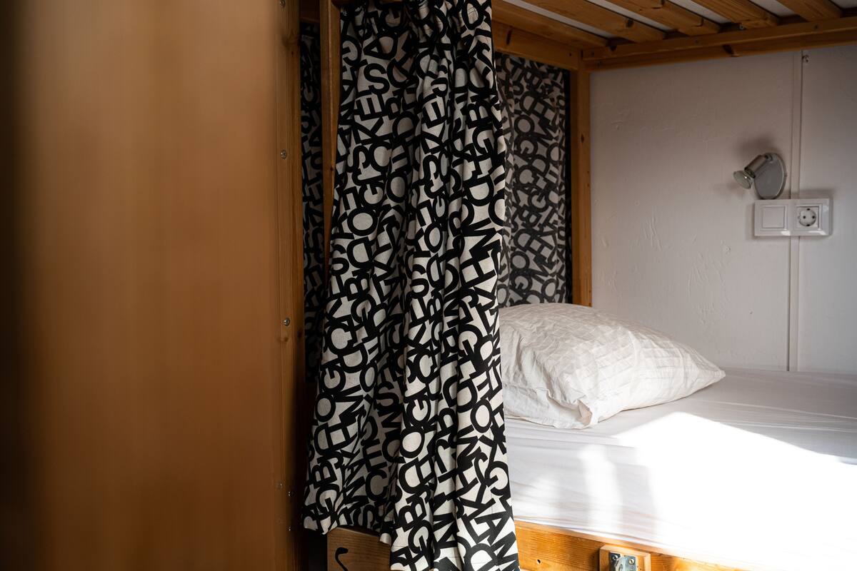 Un lit superposé avec un matelas blanc et un oreiller blanc, une tête de lit en bois et un rideau à motifs.
