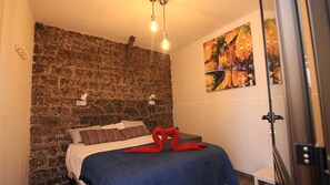 Ein Schlafzimmer mit Steinwand, einem Bett mit gestreiften Kissen, einer blauen Decke und zwei roten Schwänen auf dem Bett. An der Wand hängt ein Gemälde und zur Rechten befindet sich eine Tür.