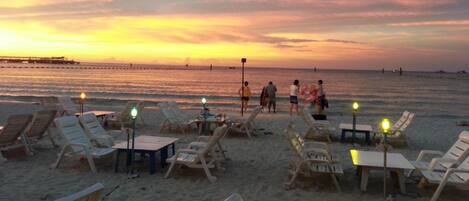 Un entorno playero con tumbonas y mesas, un muelle al fondo y una puesta de sol.