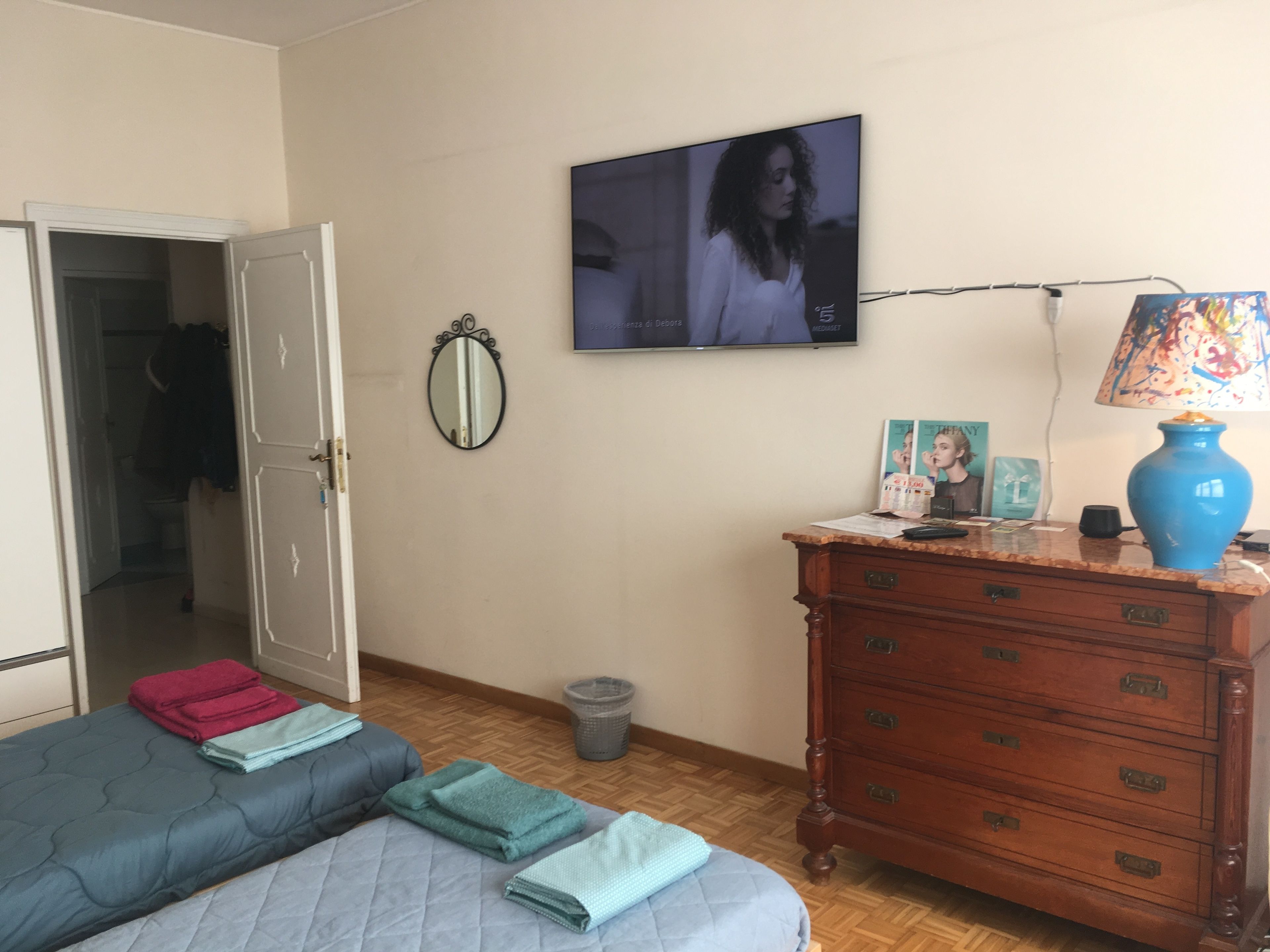 Quarto com cama, cômoda, espelho, televisão e lixeira.