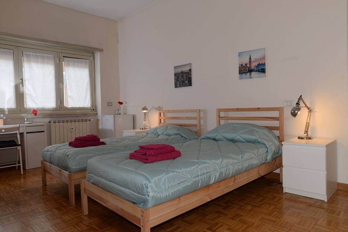 A bedroom with two beds, a desk, a radiator, and a window with curtains.