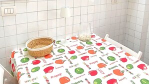 Un pequeño comedor con una mesa cubierta por un mantel con motivos de frutas, un reloj en la pared y una imagen de un paisaje urbano.