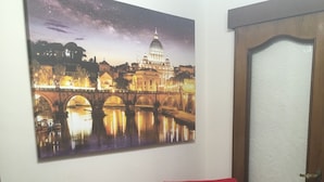 Una habitación con una silla roja, un cuadro de un paisaje urbano colgado en la pared, una puerta y un estante pequeño con revistas y un periódico.