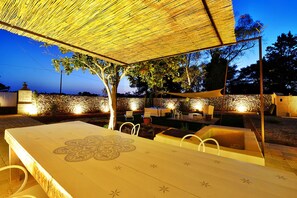 Un área de comedor al aire libre con una mesa y sillas bajo un toldo sombreado, rodeada de muros de piedra y árboles.