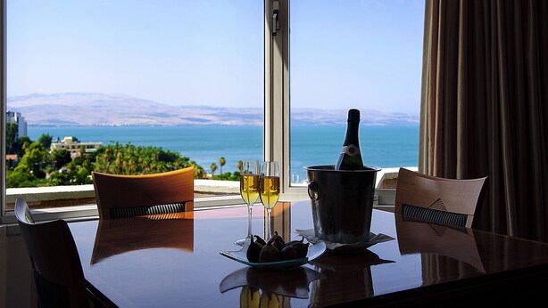 A table with a bottle of champagne, two glasses of wine, and a bowl of fruit in a hotel room with a view of the sea and mountains. - Kings Towers Suite Apartments (Tiberias)