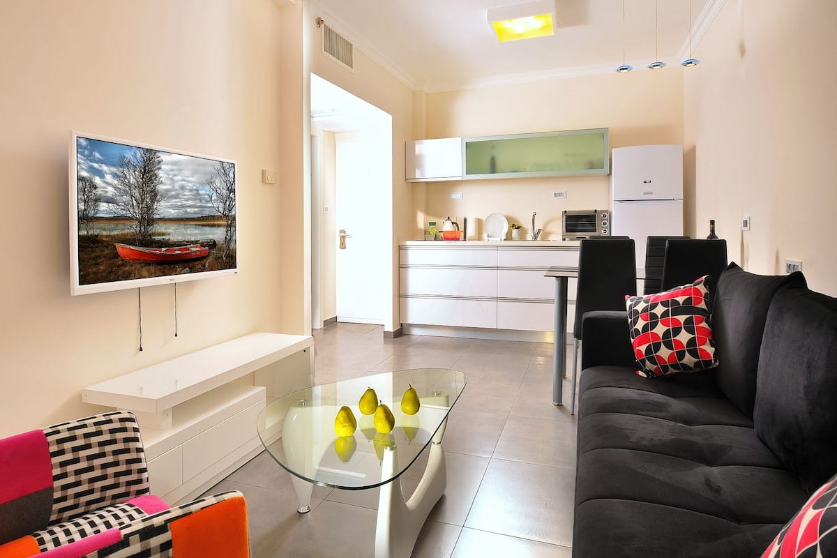 Un salon moderne avec une table basse en verre, un canapé noir agrémenté d’un coussin à motifs et une chaise colorée. On aperçoit une cuisine à l’arrière-plan, avec des meubles blancs et un four à micro-ondes.