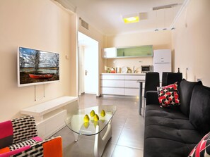 Ein modernes Wohnzimmer mit einem Glastisch, einem schwarzen Sofa mit einem gemusterten Kissen und einem farbenfrohen Sessel. Im Hintergrund ist eine Küche mit weißen Schränken und einer Mikrowelle erkennbar.