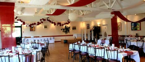 A banquet hall with tables set for a formal event, featuring white and red drapery, chandeliers, and a wooden floor.