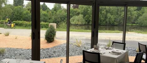A restaurant with a view of a lake and greenery. Tables are set with white tablecloths and silverware.