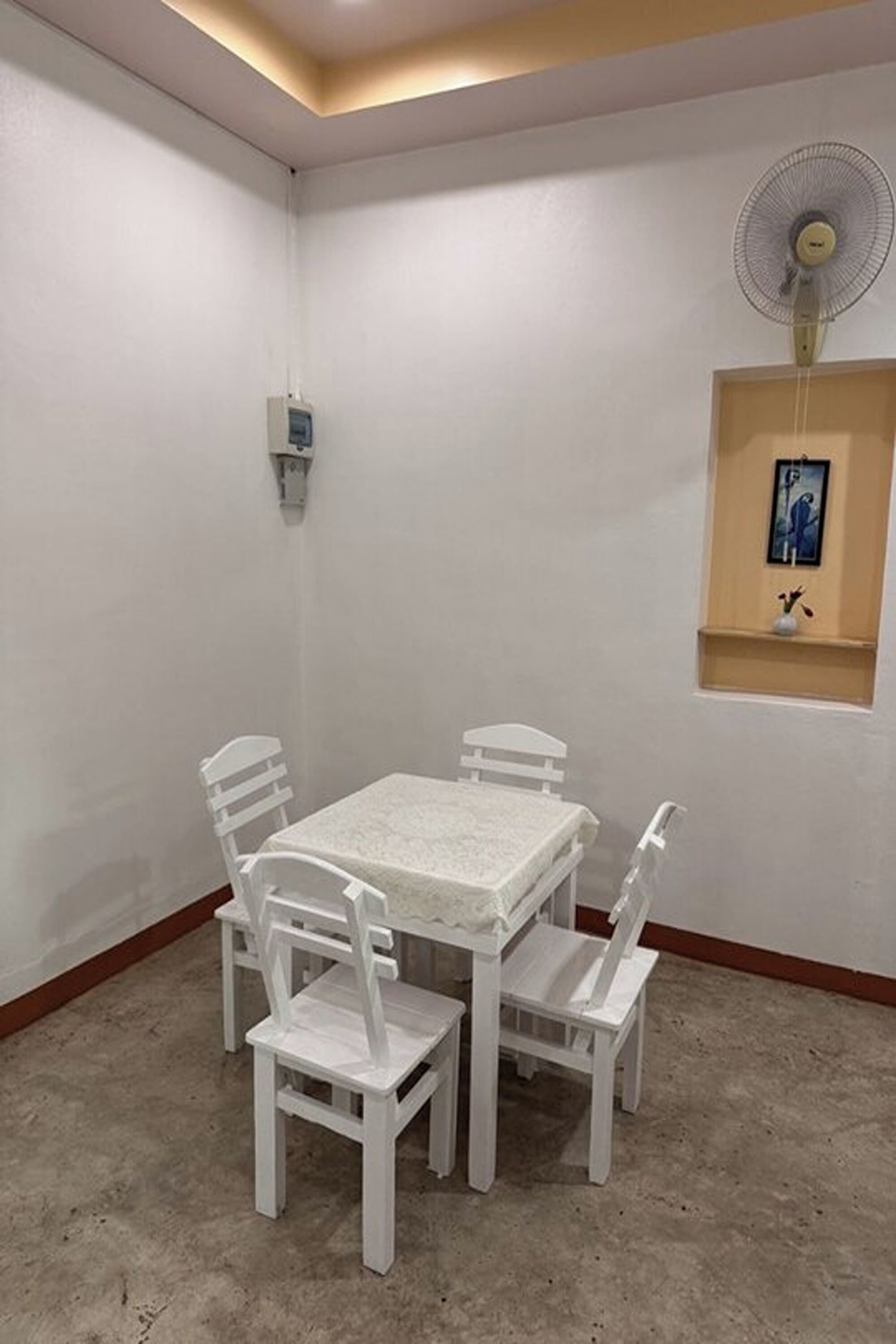 A small, white-walled room with a table and four chairs, a wall-mounted fan, and a small shelf with a picture and a vase.