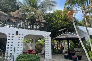 A covered outdoor seating area with tables and chairs, surrounded by palm trees and a white building with a lattice design. - Ban Suan Suree Resort (Lamphun)