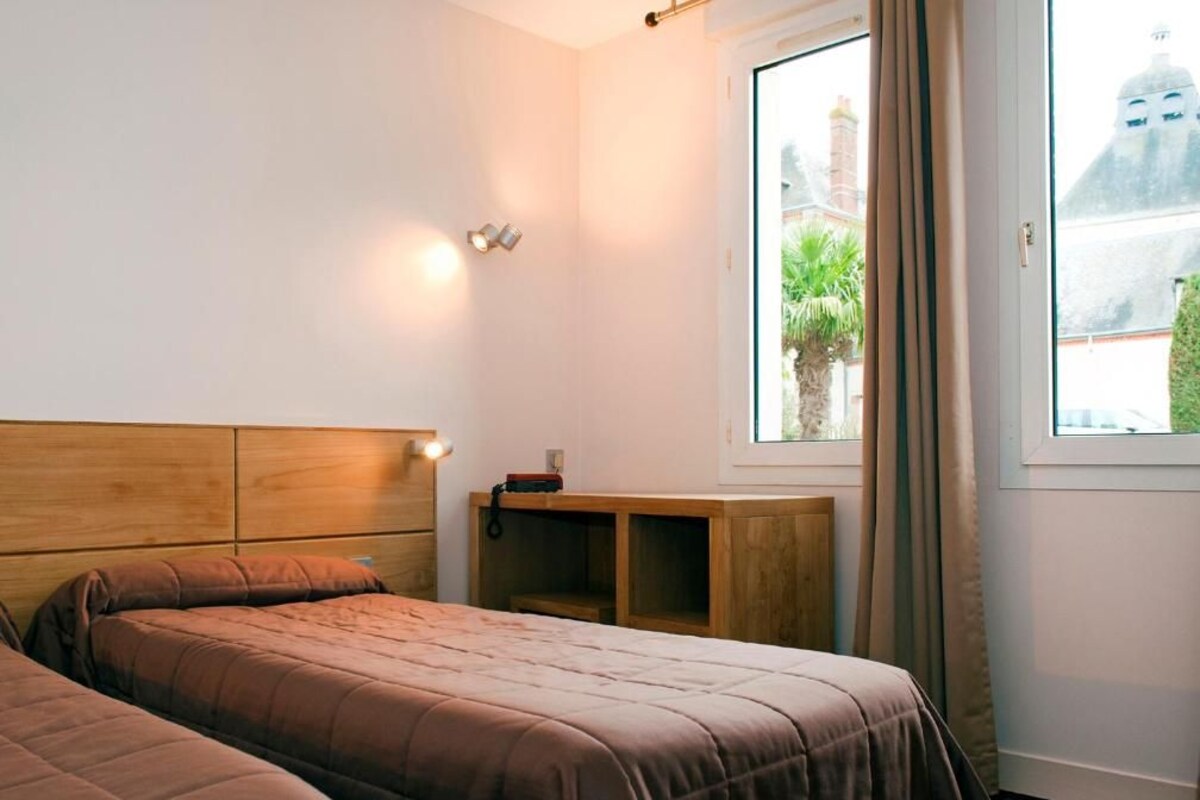 Quarto com duas camas, cabeceira de madeira, mesa de cabeceira, janela com vista para edifícios e uma porta.