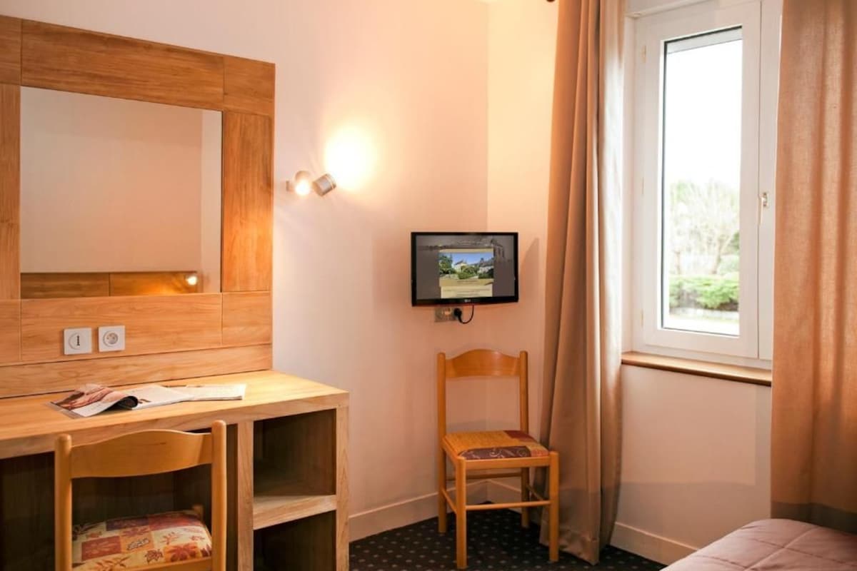 Quarto de hotel com escrivaninha de madeira, televisão, cadeira, cama e janela com cortinas.