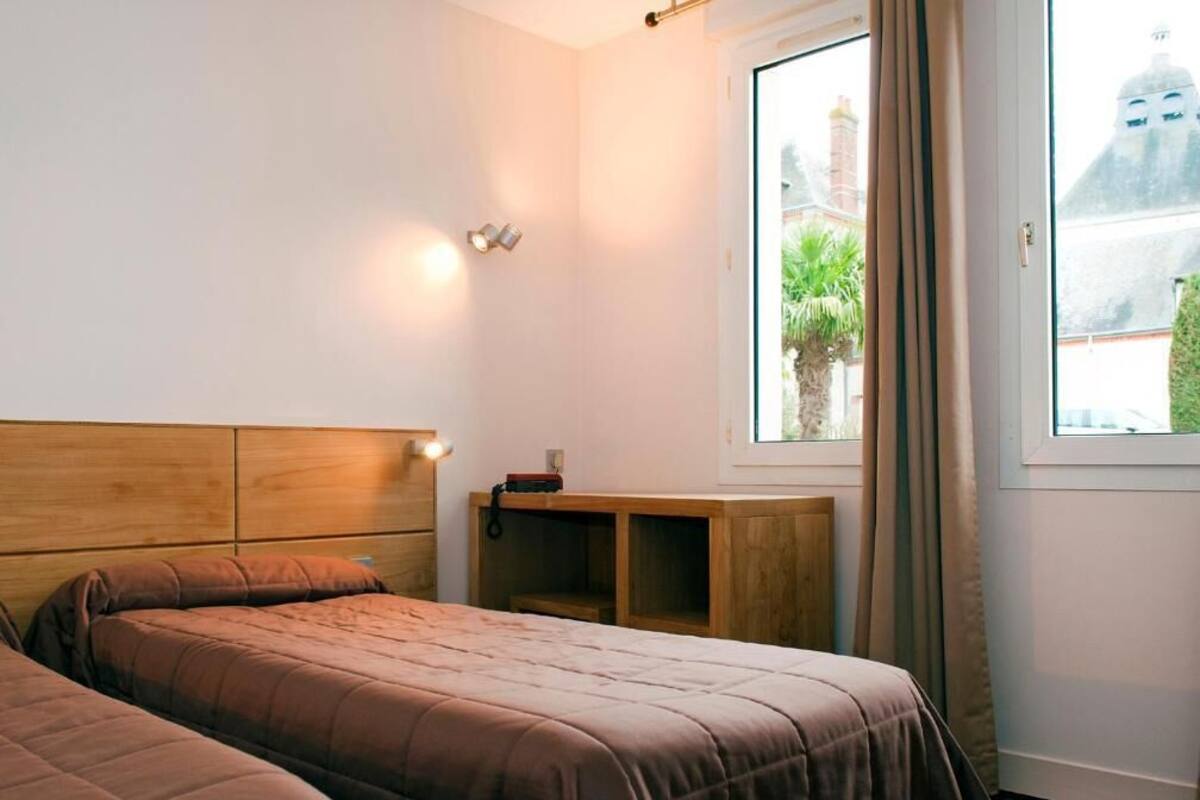A bedroom with two beds, a wooden headboard, a nightstand, a window with a view of buildings, and a door.
