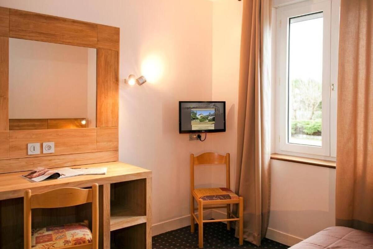 A hotel room with a wooden desk, a television, a chair, a bed, and a window with curtains.
