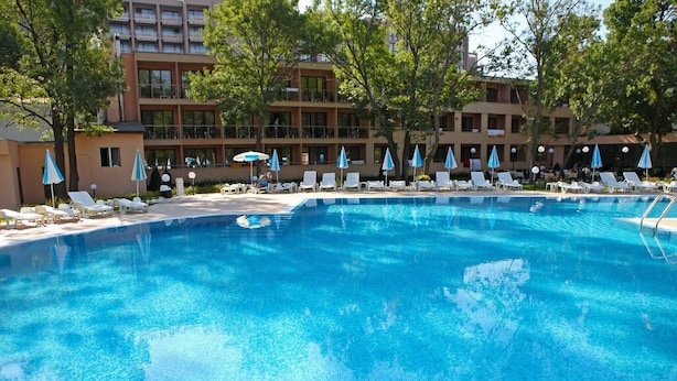 Piscina con sillones de descanso y sombrillas, rodeada de árboles y un edificio con balcones.