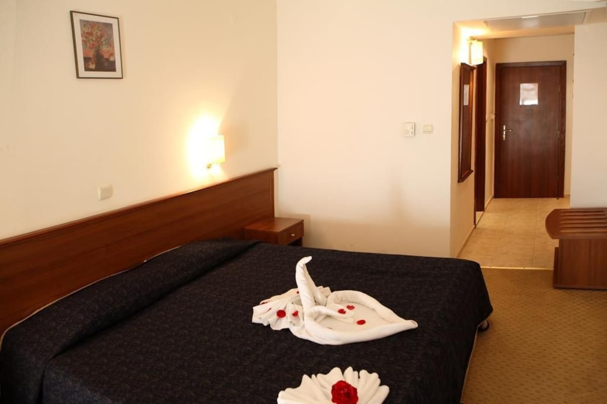 A hotel room with a neatly made bed, a bedside table, a lamp, and a door leading to another room.
