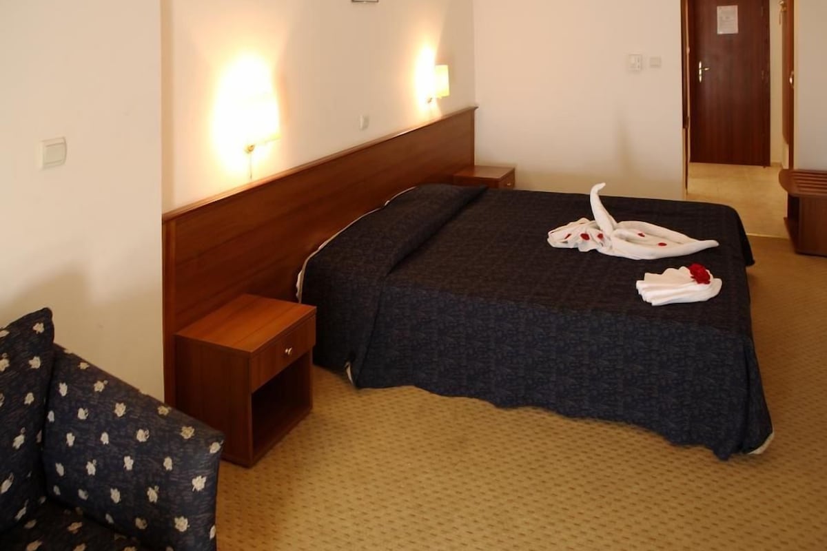 A hotel room with a large bed, a wooden nightstand, a chair, and a wall-mounted lamp.